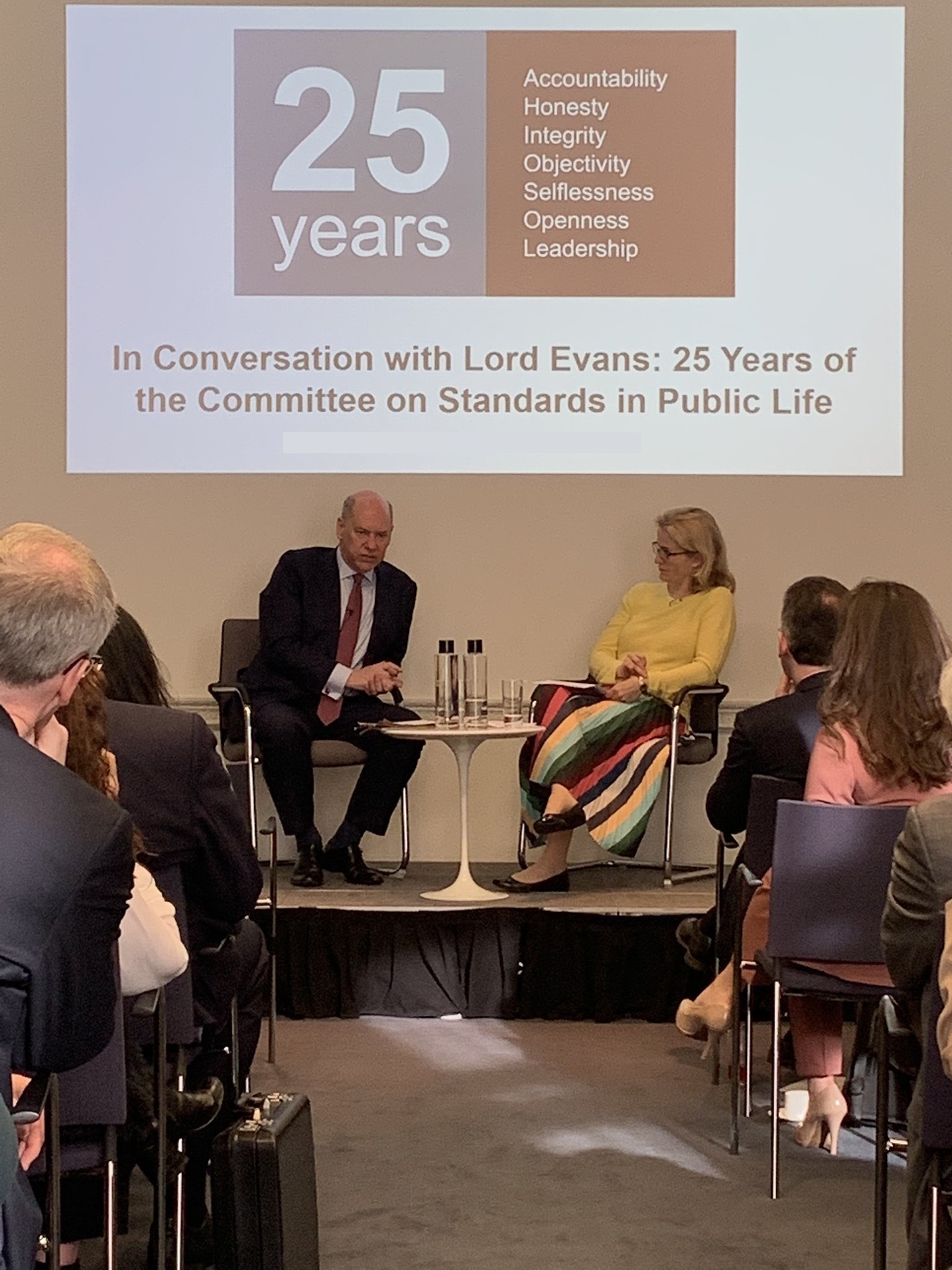 Lord Evans speaking to Hannah White in front of an audience at the IfG underneath a CSPL 25 years logo projected on the wall